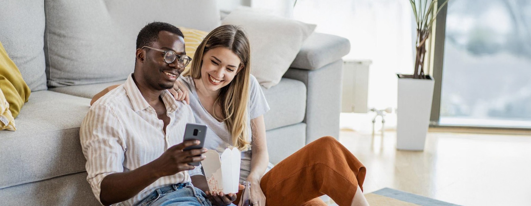 a man and woman with take out, sit against a couch on their living room floor and watch at their cell phone