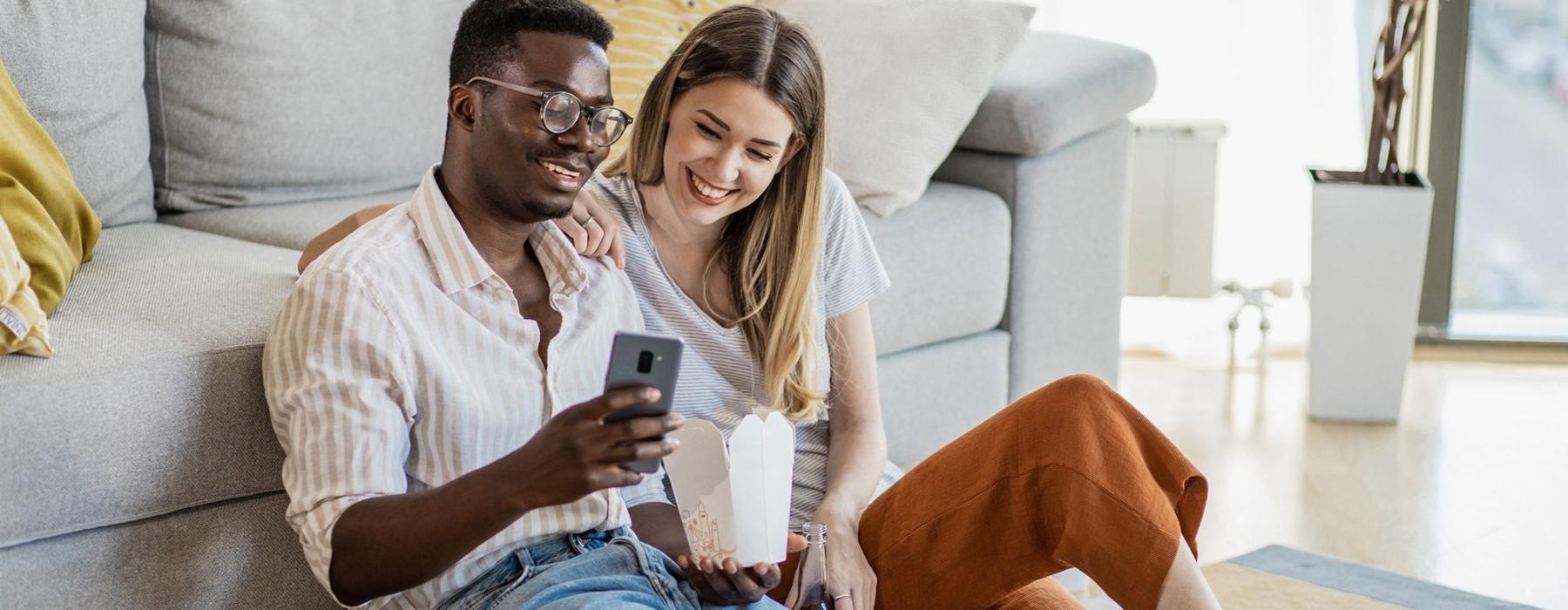 a man and woman with take out, sit against a couch on their living room floor and watch at their cell phone